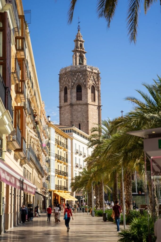 A vibrant street scene with the historic bell tower in Valencia, Spain, filled with walking pedestrians and majestic palm trees.